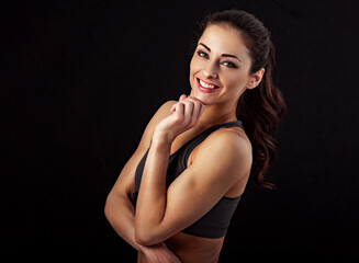 Fototapeta na wymiar Confident sport woman in sport bra smiling and looking in camera, fitness trainer standing front view, workout in gym isolated on black background with empty copy space. Closeup