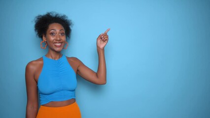 Happy African-American young woman in blue clothes posing on blue background and smiling, pointing with finger at empty space aside her, shopping time concept, copy space