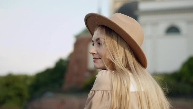 Portrait happy young caucasian woman with long blonde hair wearing fedora hat stands in front of building in historical part of city on sunny day. Cute female looking at cityscape and smiles to camera