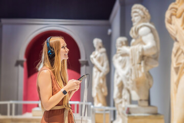 Portrait of contemporary young woman looking at sculptures and listening to audio guide at museum...