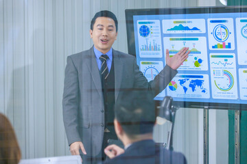 Millennial Asian professional successful male businessman lecturer presenter in formal business suit standing pointing computer monitor presenting graph chart financial info to businessman client