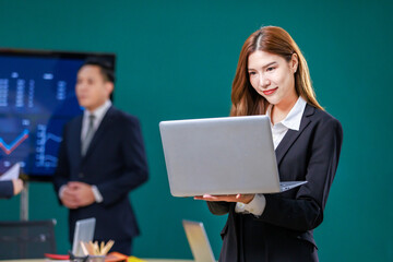 Fototapeta na wymiar Millennial Asian professional successful businesswoman in formal business suit standing holding laptop computer showing fist up celebrating working goal achievement in meeting room with colleagues