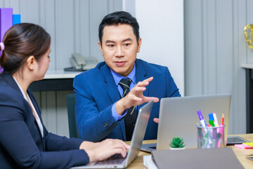 Millennial Asian professional successful male businessman in formal business suit sitting talking discussing with female businesswoman colleague typing laptop computer in company office workstation