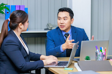 Millennial Asian professional successful male businessman in formal business suit sitting talking discussing with female businesswoman colleague typing laptop computer in company office workstation