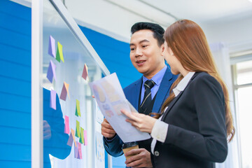 Asian professional successful male businessman in formal business suit standing reading pointing discussing memo data on post it sticky note on glass board and paperwork with female businesswoman