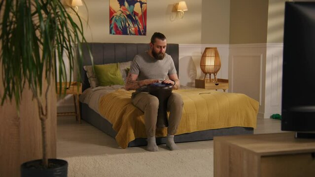 Concentrated man sits on the bed in bedroom and rhythmic plays on traditional djembe drum. Musician practices skills in cozy home. House with minimalist modern design. Music training concept. Zoom in.