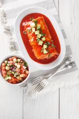 Mexican wet burrito platter with red enchilada sauce, refried beans, rice and cheese closeup on the plate on the wooden table. Vertical top view from above