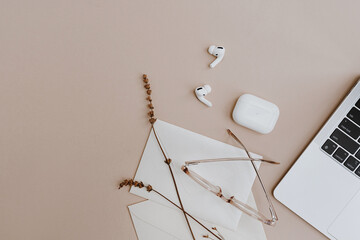 Stylish office workspace desk. Wireless earphones, glasses, laptop, envelopes on tan beige table. Aesthetic neutral tan beige and white colours.
