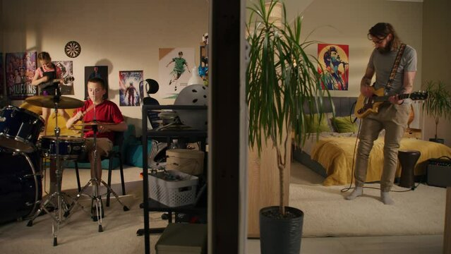 Man plays guitar in bedroom. Young boy plays drum kit and young girl plays ukulele, jumps on bed. View of two rooms or apartments separated by wall. Concept of music, neighbourhood and lifestyle.