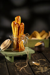 Churros served in a glass jar with caramel sauce, placed on a rustic wooden surface. Crispy golden pastry sticks, sugar crumbs, and warm tones create a cozy dessert atmosphere.