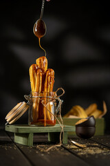 Churros served in a glass jar with caramel sauce, placed on a rustic wooden surface. Crispy golden pastry sticks, sugar crumbs, and warm tones create a cozy dessert atmosphere.