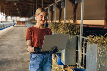 A beautiful Caucasian woman is standing n a stable with a laptop.