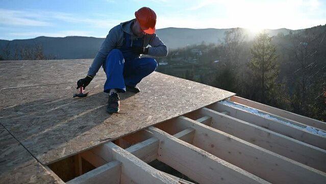 Carpenter hammering nail into OSB panel on the roof top of future cottage at sunset. Man worker building wooden frame house. Carpentry and construction concept.