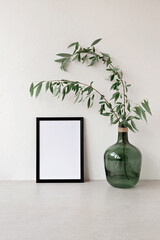 Empty black picture frame mockup and green glass jar with tree branches on neutral beige table, concrete wall background. Modern interior decoration, aesthetic minimal art poster template, copy space
