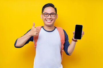 Cheerful young Asian man student in casual clothes backpack showing blank screen smartphone, thumb up gesture, recommends new app isolated on yellow background. high school university college concept