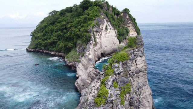 Waves breaking on cliffs of lush green limestone sea mount rock formation against Atuh Beach, Nusa Penida Island. Aerial