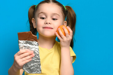 Tangerine and chocolate bar in child's hands, concept of healthy and unhealthy food on blue background.