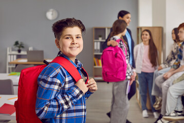 Portrait of smiling boy school student standing with backpack. Happy schoolboy wearing casual clothes posing on background of his classmates in classroom and looking at camera