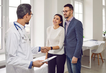 Obraz premium Young beautiful couple at doctor's consultation in the office of the Family planning medical center. Professional obstetrician-gynecologist talks to happy family, shaking hands with smiling girl.