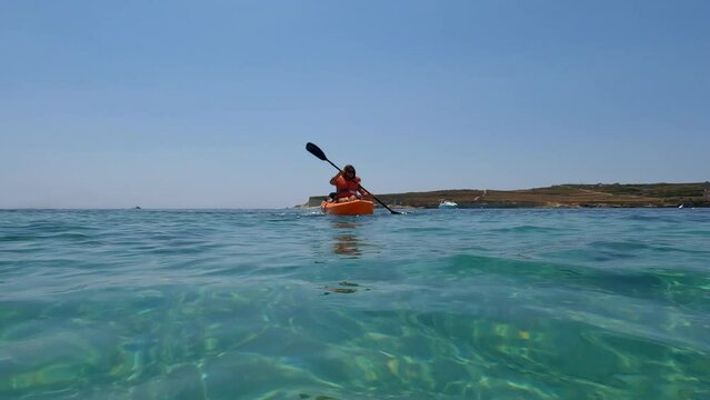 Water level view of a woman with an orange kayak passing by at sea.