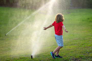 Cute little kid watering grass in the garden at summer day. Child play in summer backyard. Cute little boy is laughing and having fun running under water spraying sprinkler irrigation. Watering grass. - obrazy, fototapety, plakaty