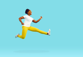Fototapeta na wymiar Happy funny young dark skinned woman having fun running and jumping on light blue background. Full length woman in white t-shirt, yellow pants and sunglasses fooling around near copy space.