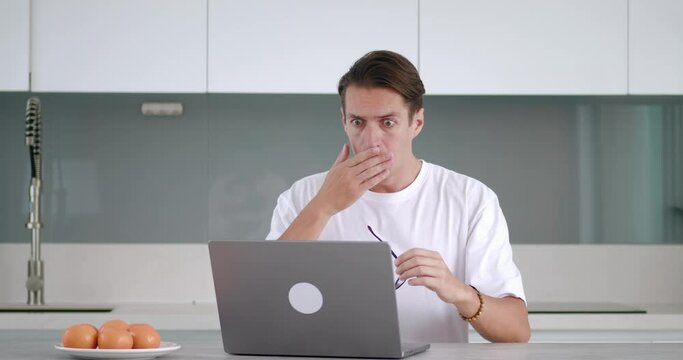Man is shocked while reading bad news at home kitchen. Stressed male in glasses cant believe at laptop working fail problems. Business man is shocked looking laptop screen, working from home