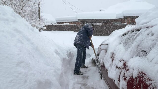 A man in a cap, jacket and jeans is shoveling snow next to his car. There are big drifts all around. In the background is a cinnamon fence and a house. Roofs are covered with a thick layer of snow