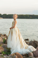 Beautiful young girl  in luxurious fluffy white dress with gold trim standing  sideways looking at camera on the bank of river or lake with big stones in the evening, romantic outdoor kid's portrait