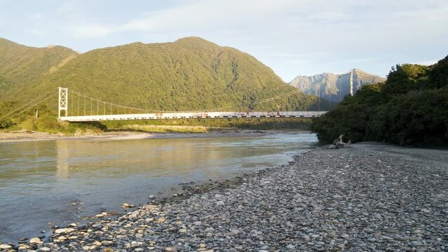 Rusty suspension bridge in mountains at sunset; low aerial orbit