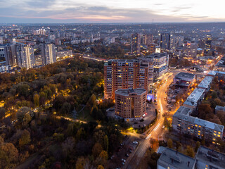 Aerial view of Kharkiv city park, Botanical garden in Sarzhyn Yar near multistory modern high buildings in evening. City with street lights illumination and driving cars