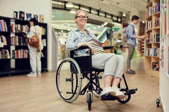 Portrait of happy smart person with disability girl with blond hair sitting in wheelchair and holding books in university library - Powered by Adobe