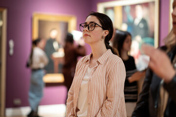 Portrait of young Caucasian woman wearing glasses and looking at exhibition. People and masterpieces in background. Cultural education. Concept of Museum Day