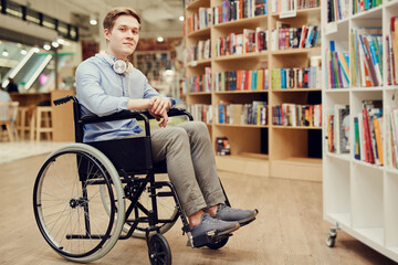 Portrait of content student boy with paralyzed legs sitting in wheelchair and choosing books in library