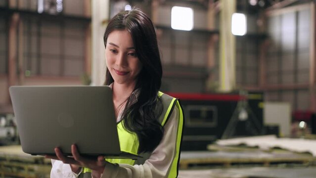 Asian Engineer woman wear vest safety typing laptop computer while checking details of product inventory in warehouse at steel part factory