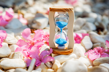 hourglass with blue sand on white stones and pink flowers, time passing very fast concept
