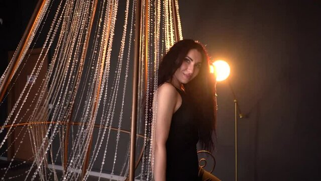 A beautiful brunette in a black suit looks at the camera through the light and glare against the background of a crystal chandelier in a dark studio