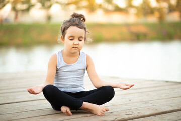 Llittle girl child kid sitting in nature near by river or lake and practicing yoga. Healthy lifestyle.