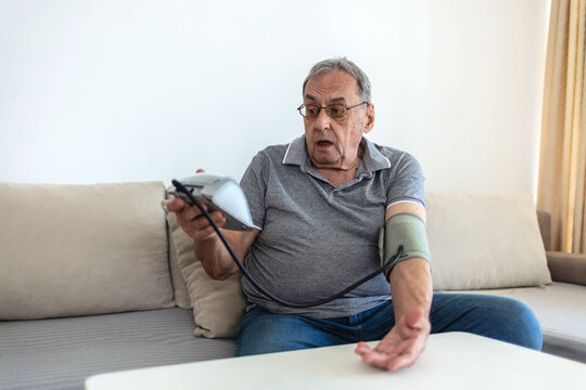 Senior man sitting in his living room on a sofa and checking his blood pressure. Photo of mature man measuring her blood pressure at home.