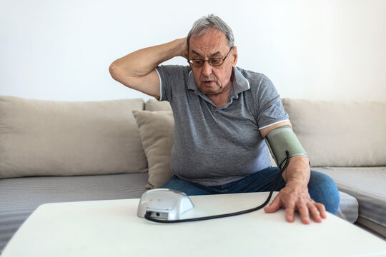 Senior man sitting in his living room on a sofa and checking his blood pressure. Blood pressure monitor - man using in home sitting on sofa.