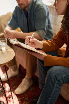 Vertical image of therapist talking to patient and making notes in notepad while they sitting on sofa in office