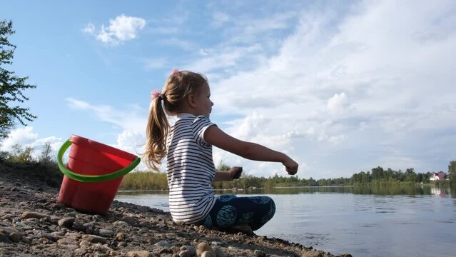 Child girl playing on the beach and throwing  stones into the river. Investigating details of nature . 
