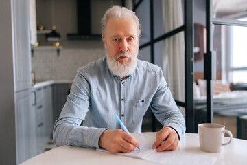 Portrait of confident bearded senior older man writing making notes in notebook, keep personal...