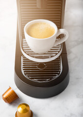 White coffee cup and home espresso machine with coffee capsules on white marble kitchen background.