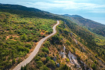 Aerial view of mountain road, blue sky with clouds at  summer sunny day. Lefkada, Greece. Top view from drone of empty road, green forest on hills in spring. Beautiful landscape with highway. Travel