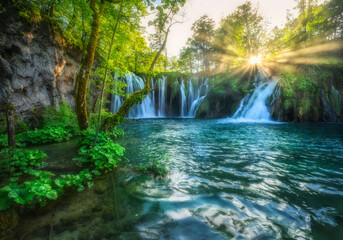 Fototapeta na wymiar Waterfall in green forest in Plitvice Lakes, Croatia at sunset in summer. Colorful landscape with fall, blooming park, trees, water lilies, sunbeams, river in spring. Scenery. Park in woods. Nature 