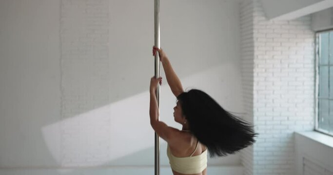 Smiling Asian strip dancer spinning on a pylon, in bright white studio in daylight against the background of a panoramic window in beige lingerie. Flexible gorgeous stripper goes up and down the pole