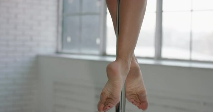 Asian strip dancer. Woman's feet on a pole with red pedicure. Pole dance woman twirling on pole bright studio Panoramic windows background. Slow motion. Beautiful female ankles knees rotate on a pole