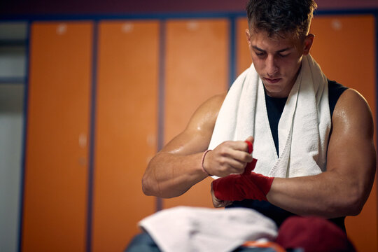 Wide horizontal shot of young sweaty man taking red protective tape of his hands after boxing workout.