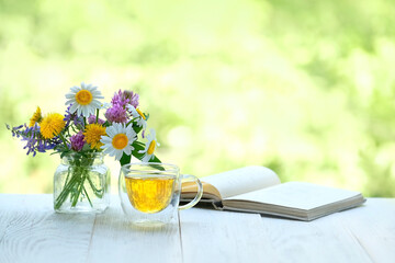 Beautiful meadow flowers bouquet, glass cup with herbal tea and book close up on table in garden,...